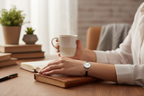 Person holding a cup of coffee with a watch on a wooden table, surrounded by books and a plant.