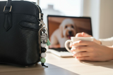 Black handbag with a keychain on a desk next to a laptop displaying a dog, with a person holding a mug.