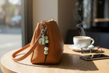 Brown leather handbag with keychain on a wooden table next to a steaming cup of coffee.