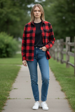 Woman wearing a red and black plaid shirt, black top, blue jeans, and white sneakers standing on a path with greenery in the background.